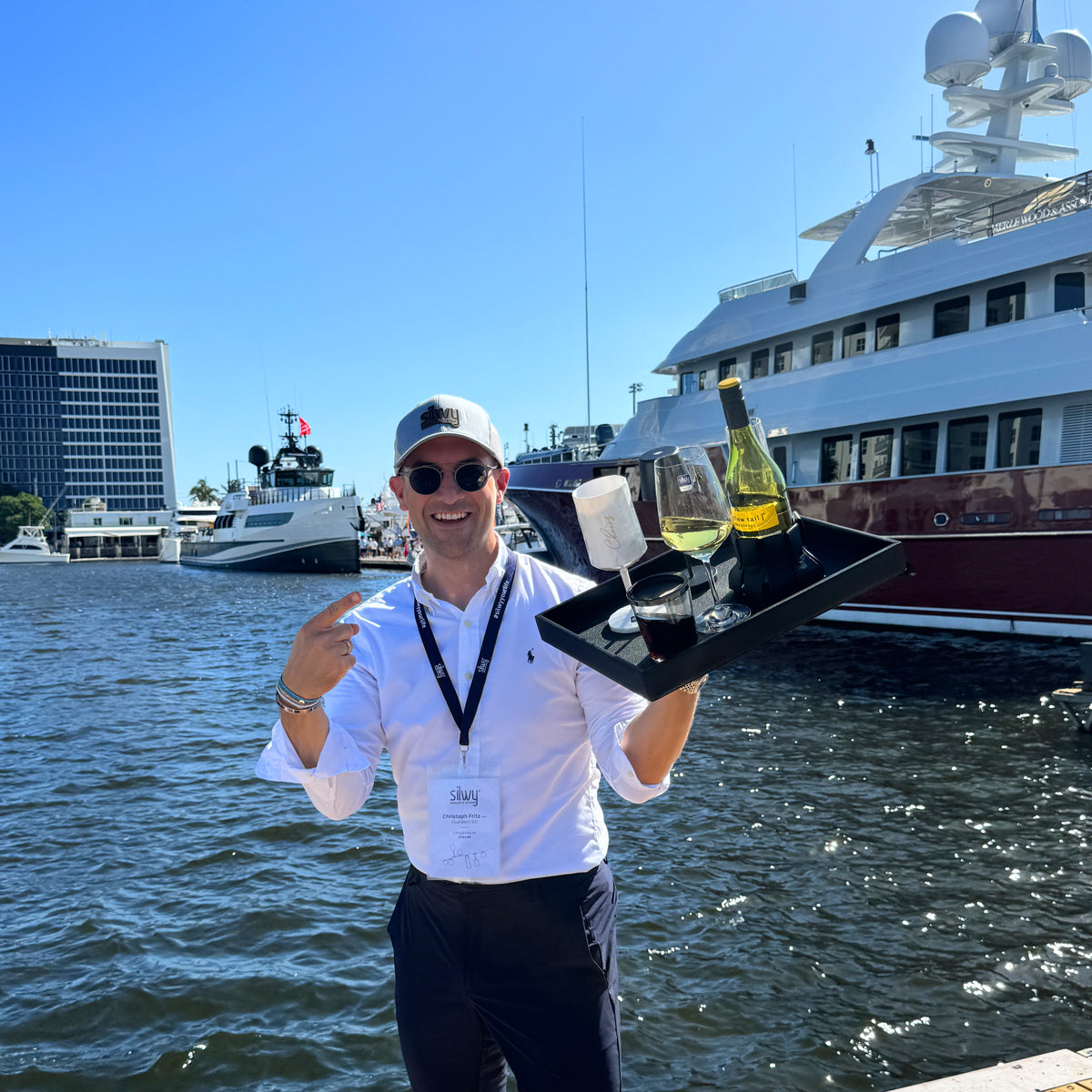 Founder und CEO Christoph mit einem Metall-Tablett in der Hand. Darauf befindet sich ein Magnet-Kristallglas WEIN, einem Magnet-Kunststoffglas WEIN CHEERS in weiß, einem Magnet-Trinkbecher Triple Classic und eine Flasche Wein. Im Hintergrund ist ein Yachthafen mit Schiffen zu sehen 